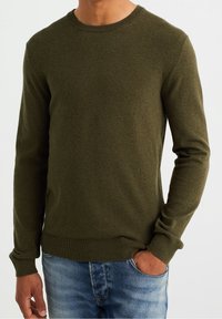 Olivově zelený pletený svetr s kulatým výstřihem a dlouhými rukávy. Hladké manžety a lem, texturovaná látka, spárováno s modrými džíny.