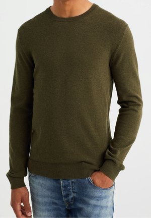 Olivgrüner Strickpullover mit Rundhalsausschnitt und langen Ärmeln. Gerippte Bündchen und Saum, strukturierter Stoff, kombiniert mit blauen Jeans.