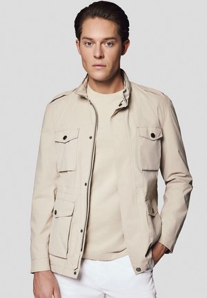 Jasnobeżowa kurtka z zamkiem z przodu i dwoma kieszeniami na piersi, z wyróżniającym się kołnierzem i akcentami w postaci guzików. Noszona na beżowej koszuli.