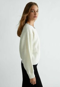 Maglione bianco lavorato a maglia con una trama a coste, caratterizzato da un contorno decorato e un colletto ornamentale. Abbinato a una gonna nera.