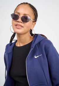 Tummansininen vetoketjullinen huppari, jossa on valkoinen Nike-logo, päällä musta ribattu toppi. Mallilla on tummat soikeat aurinkolasit ja suuret hopeiset korvaketjut.