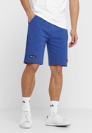 Homme portant un short bleu ellesse, des chaussettes blanches FILA, des baskets blanches et un t-shirt blanc, debout avec une main dans la poche.