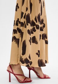 Voeten in rode sandalen met hoge hakken en banden onder een beigekleurige rok met grote donkeren bruine luipaardvlekken.