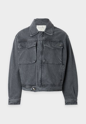 Uomo che indossa una giacca di jeans grigia sbiadita, pantaloni neri larghi e scarpe beige, in piedi su uno sfondo bianco semplice.