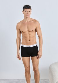Black boxer briefs with a white TOMMY HILFIGER logo elastic waistband, fitted design, and smooth fabric. Model stands against a light background.