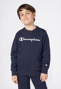 Ragazzo che indossa una felpa Champion blu navy e pantaloni abbinati, in piedi con un leggero sorriso su uno sfondo semplice.