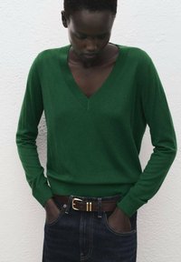 Maglione verde a scollo a V realizzato in morbido materiale lavorato a maglia, con maniche lunghe e polsini a coste. Abbinato a una cintura marrone e jeans scuri.