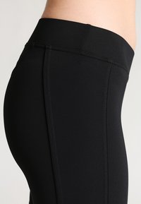 Primo piano di una persona che indossa leggings neri a vita alta con dettagli delle cuciture in evidenza su uno sfondo neutro.
