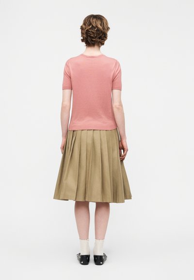 Γυναίκα με κοντά σγουρά μαλλιά που φοράει ροζ μπλουζάκι με κοντά μανίκια, μπεζ πλισέ φούστα μέχρι το γόνατο, λευκές κάλτσες και μαύρα παπούτσια, κοιτάζοντας προς τα πίσω.