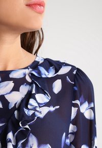 Bluse i mørk marineblå stof med blomsterprint, der har flæsekant ved halsen, stort mønster med hvide og blå kronblade samt plisserede, puffede ærmer.