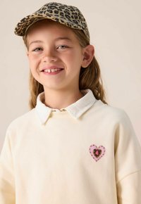 Crème sweatshirt met een witte kraag, voorzien van een roze hartpatch met een luipaardprint binnenin. De persoon draagt een pet met luipaardprint.