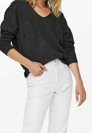 Femme portant un pull à col en V gris foncé et un pantalon blanc taille haute, avec des bagues aux deux mains, sur fond blanc.