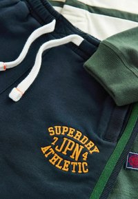 Navy and green sweatshirt with white drawstrings, featuring orange embroidered text "SUPERDRY 7JPN 4 ATHLETIC" and textured cuffs.