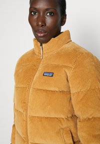 Chaqueta de abrigo de pana color mostaza con un alto cuello, cierre de cremallera y parche del logo azul de Patagonia en el pecho. Tela texturizada y acanalada.