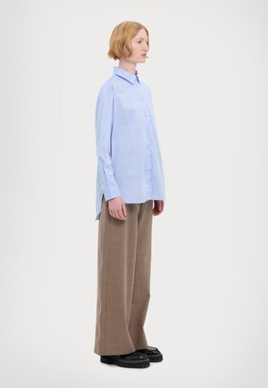 Light blue button-up shirt with long sleeves, featuring a side split; paired with loose, brown plaid trousers and black platform shoes.