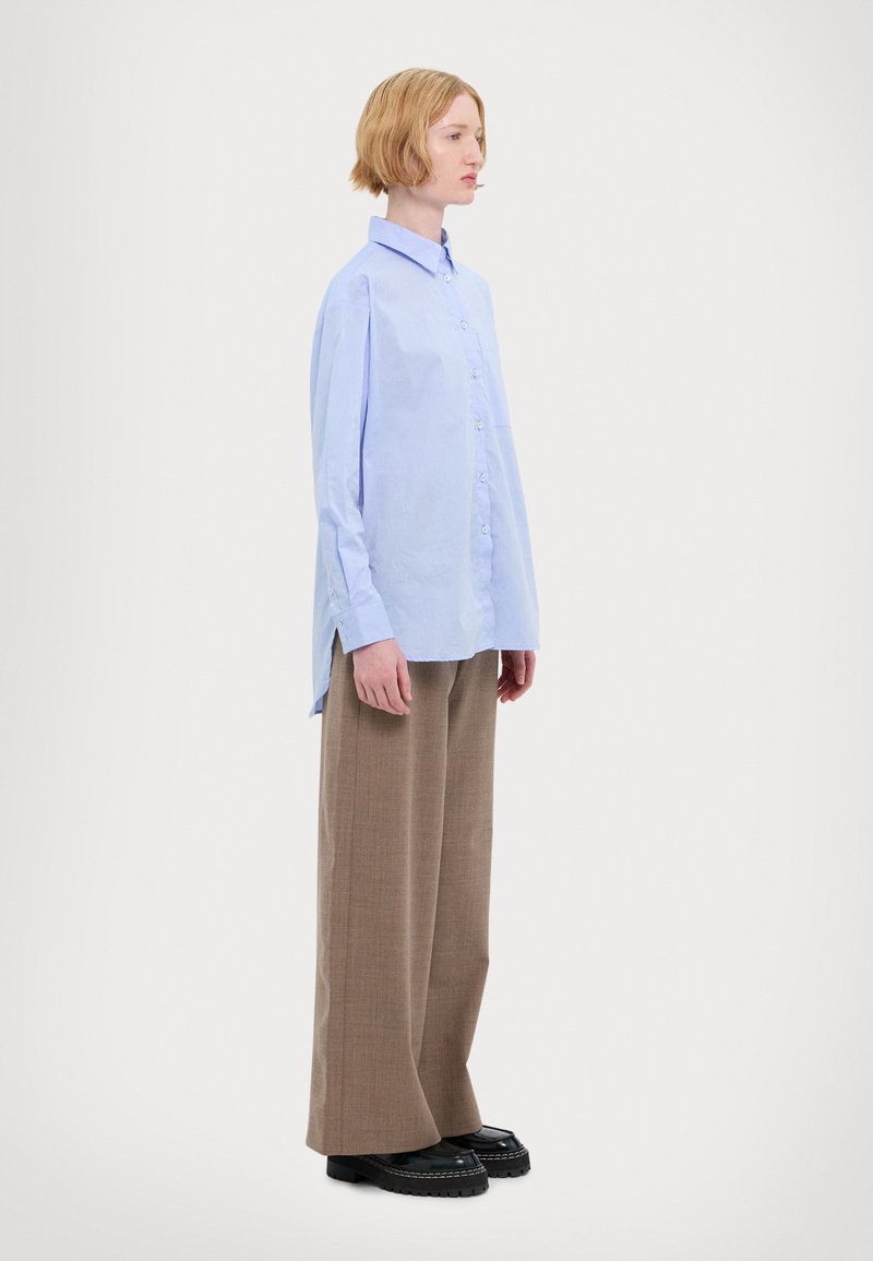 Camicia button-up azzurro chiaro con maniche lunghe, caratterizzata da uno spacco laterale; abbinata a pantaloni a quadri marroni larghi e scarpe con zeppa nere.