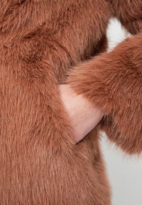 Καφέ παλτό από οικολογική γούνα με απαλή, αφράτη υφή, που διαθέτει ευρύ μανσέτα και άνετη εφαρμογή, αποκαλύπτοντας ένα χέρι που ακουμπάει στο πλάι.