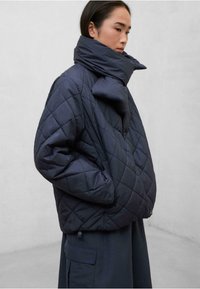 Chaqueta de lana acolchada en color azul marino con un cuello alto, que presenta un distintivo detalle de lazo en el cuello y bolsillos laterales, fabricada con una tela texturizada.