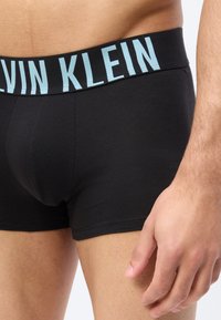 Άνδρας μέσης και άνω μηρού που φοράει μαύρα ανδρικά εσώρουχα Calvin Klein με ανοιχτό μπλε λογότυπο στη ζώνη, το δεξί χέρι χαλαρό στο πλάι.