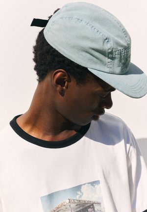 Mladý muž so svetlomodrou čiapkou a bielym tričkom s čiernym golierom a grafickou potlačou sa pozerá dole na svoju pravú stranu.