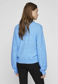 Svetlo modra pletena jopica z ohlapnim krojem, ki ima očesne vzorce in rebraste manšete ter rob. Gladka tekstura s sproščenim silhuetom.