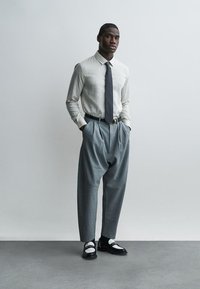 Άνδρας στέκεται με τα χέρια στις τσέπες, φοράει ριγέ πουκάμισο με μακριά μανίκια, σκούρα γραβάτα, γκρι ψηλόμεσα παντελόνια με πιέτες και ασπρόμαυρα παπούτσια.