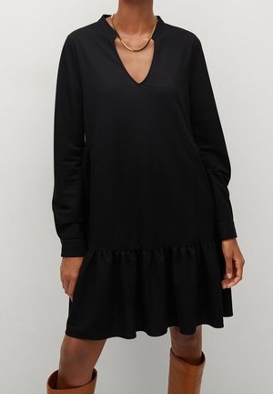 Frau trägt ein lockeres, schwarzes, langärmeliges Kleid mit V-Ausschnitt und Rüschensaum, kombiniert mit kniehohen braunen Stiefeln und einer goldenen Halskette.