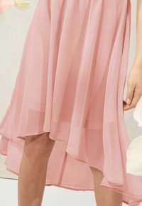 Abito rosa semi-trasparente con orlo asimmetrico e fluente. Tessuto leggero e testurizzato che si drappeggia dolcemente, creando un movimento leggero. Accentuato da morbidi pieghe.