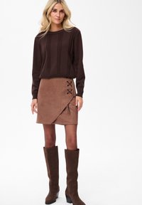 Blonde vrouw in een donkerbruin kabelgebreid vest, een tan suède wikkelrok met zwarte vetersluiting en bruin kniehoge laarzen.