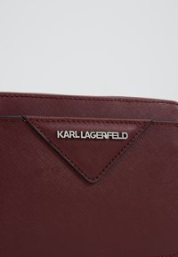 Burgundová kožená peněženka s texturovaným povrchem, zdobená stříbrným logem "KARL LAGERFELD" a trojúhelníkovým dekoračním prvkem.