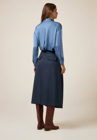 Donna in piedi di lato che indossa una blusa blu a maniche lunghe, una gonna blu scura di lunghezza media e stivali marroni fino al ginocchio su uno sfondo semplice.