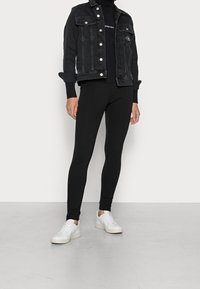 Veste en jean noire avec boutons argentés portée sur un col roulé noir et des leggings noirs. Le modèle porte des baskets blanches au design minimaliste.