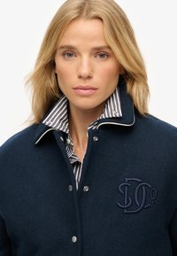 Abrigo de lana azul marino con un cuello alto, botones a presión plateados y un logo bordado en azul oscuro. Se ve una camisa a rayas debajo.