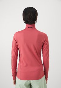 Persona con cabello corto y oscuro, vistiendo una camiseta ajustada de manga larga rosa y pantalones verde claro, mostrada de espaldas contra un fondo liso.