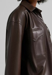 Brun läderskjorta med krage, knapplist och bröstficka; slät textur med en subtil glans och skräddarsydd passform.