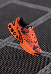 Bright orange and black athletic shoe with air cushioning and patterned design on a metal grate surface.