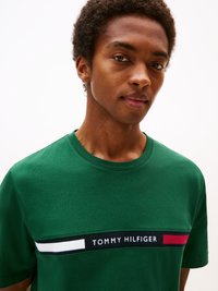 Camiseta de algodón verde con cuello redondo y bandas horizontales en blanco, rojo y azul marino en contraste, que presenta el texto "TOMMY HILFIGER" en el pecho.