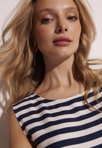 Granatowa i biała bluzka w paski z okrągłym dekoltem, wykonana z gładkiego materiału i z krótkimi rękawami. Modelka ma długie, faliste włosy.