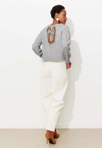 Pull tricot gris avec une ouverture au dos nouée en un nœud, associé à un pantalon en velours côtelé couleur crème et des talons marron. Texture lisse et coupe décontractée.