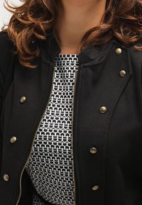 Czarna bomberka z zamkiem i mosiężnymi zatrzaskami, noszona na wzorzystej czarno-białej sukience o geometrycznym wzorze.