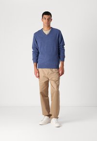 Niebieski wełniany sweter w serek nałożony na jasnoszary T-shirt, zestawiony z beżowymi spodniami i białymi trampkami, stojący na jednolitym tle.