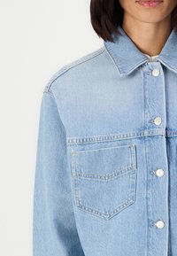 Giacca di jeans azzurro chiaro con colletto classico, bottoni frontali e una tasca sul petto caratterizzata da un distintivo design triangolare.