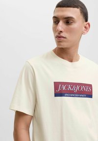Bežinė medvilninė marškinė su apvaliu iškirpimu. Priekyje yra stačiakampis grafikas tamsiai raudonos ir jūros mėlynos spalvų su užrašu "JACK & JONES" baltu tekstu.