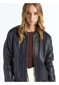 Zwarte leren bomberjack met een ritssluiting aan de voorkant, geribbelde manchetten en een aangesloten taille. Gladde textuur, oversized mouwen en minimale details.