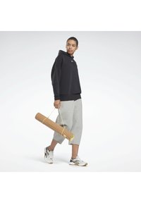 Schwarzer Kapuzenpullover mit lockerem Schnitt, graue, verkürzte Sweatpants und schwarz-weiße Sneakers. Hält eine gerollte Kork-Yogamatte mit gelbem Tragegurt.