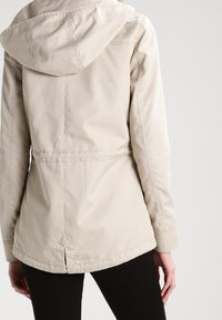 Veste imperméable beige avec capuche, silhouette ajustée, taille cintrée et coutures apparentes. Comprend un ourlet incurvé et une fente au dos.