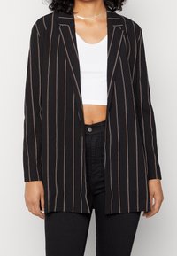 Svart blazer med vertikala vita och orange ränder, med en lång, öppen design och ett slätt, smooth tyg. Stylerad över en vit topp.