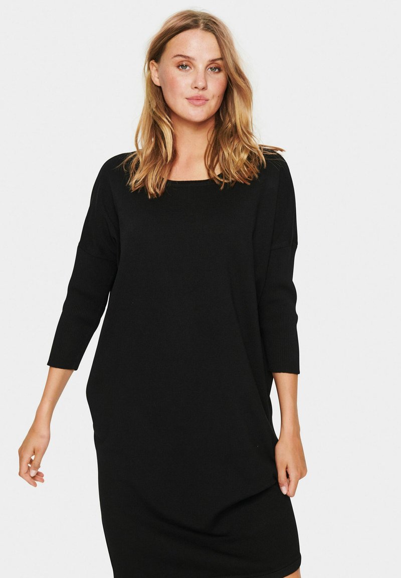 Abito nero in maglia con scollo quadrato, maniche a tre quarti e vestibilità comoda. Il tessuto ha una superficie liscia. Nessun motivo o accessorio visibile.