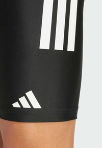 Crne sportske kratke hlače izrađene od rastezljivog materijala, s tri bijele pruge i trokutastim logotipom na donjem rubu.