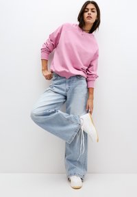 Felpa rosa chiaro con scollatura rotonda e polsini a coste, abbinata a jeans svasati azzurri e scarpe da ginnastica bianche con suola in gomma.